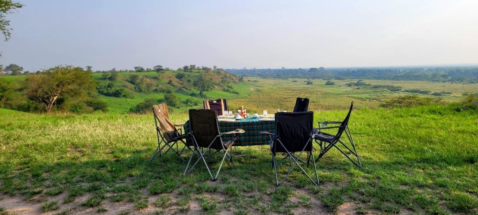 Ein gedeckter Frühstückstisch mit Campingstühlen steht im Grasland mit weitem Blick über die Savanne Ugandas